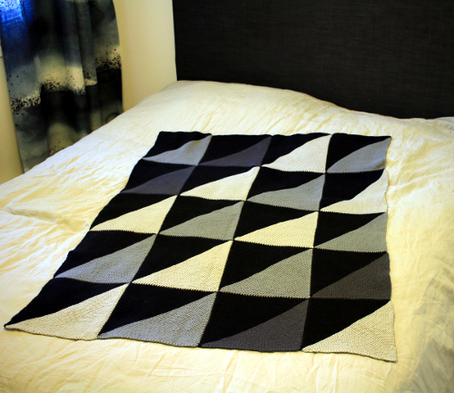 Harmaasävyisistä kolmioista koostuva Lodicula-peitto levitettynä sängylle.